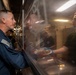 The image shows a military food service line, likely aboard a naval vessel, where personnel in a 'Food Service Division' shirt are serving food to uniformed service members. A person in a blue flight suit with a U.S. flag patch is receiving a plate of food through a sneeze guard. The setting appears to be a ship's galley or mess hall.