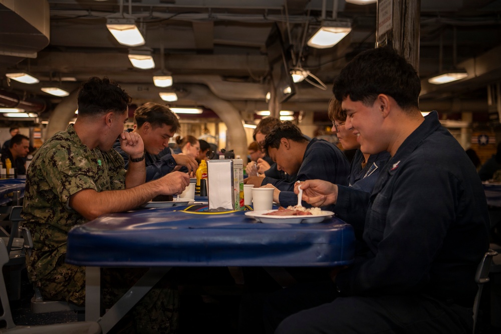 Military personnel, appearing to be U.S. Navy sailors, are eating a meal together in what appears to be a ship's mess hall or galley. The setting features industrial ceiling infrastructure, fluorescent lighting, and blue cafeteria-style tables typical of naval vessel interiors. Personnel are wearing a mix of Navy Working Uniform (NWU) camouflage and blue coveralls (utility uniforms).