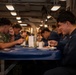 Military personnel, appearing to be U.S. Navy sailors, are eating a meal together in what appears to be a ship's mess hall or galley. The setting features industrial ceiling infrastructure, fluorescent lighting, and blue cafeteria-style tables typical of naval vessel interiors. Personnel are wearing a mix of Navy Working Uniform (NWU) camouflage and blue coveralls (utility uniforms).