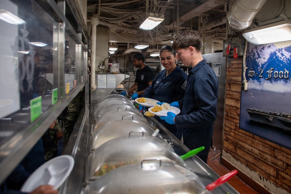 The image shows U.S. Navy personnel serving food in what appears to be a shipboard galley (mess hall). Two sailors in dark blue coveralls and blue food-service gloves are serving themselves or others from a buffet-style steam table. A sign partially visible on the right reads 'S-2 Food' alongside a display showing a naval vessel.