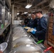 The image shows U.S. Navy personnel serving food in what appears to be a shipboard galley (mess hall). Two sailors in dark blue coveralls and blue food-service gloves are serving themselves or others from a buffet-style steam table. A sign partially visible on the right reads 'S-2 Food' alongside a display showing a naval vessel.