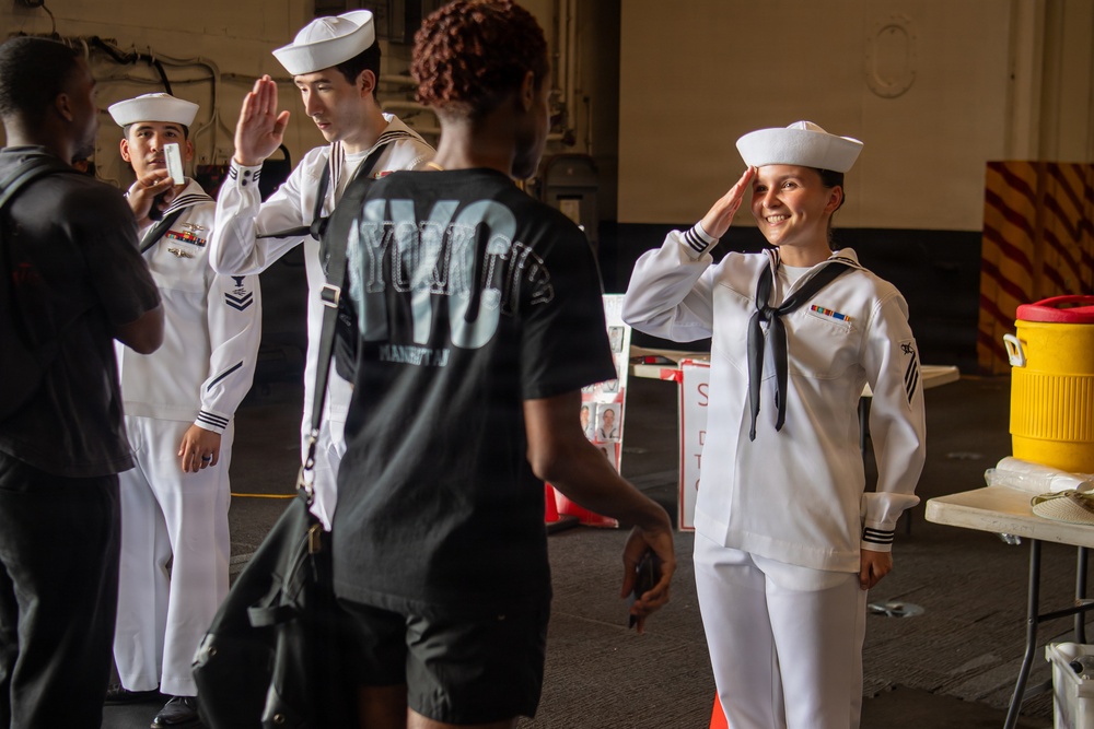 The image shows U.S. Navy sailors in white dress uniforms interacting with civilians in what appears to be a ship hangar bay or naval facility. A female sailor on the right is smiling and saluting, while a male sailor in the center is giving a high-five to a civilian wearing a black NYC jersey. The setting suggests a public outreach event, possibly Fleet Week or a ship tour.