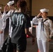 The image shows U.S. Navy sailors in white dress uniforms interacting with civilians in what appears to be a ship hangar bay or naval facility. A female sailor on the right is smiling and saluting, while a male sailor in the center is giving a high-five to a civilian wearing a black NYC jersey. The setting suggests a public outreach event, possibly Fleet Week or a ship tour.
