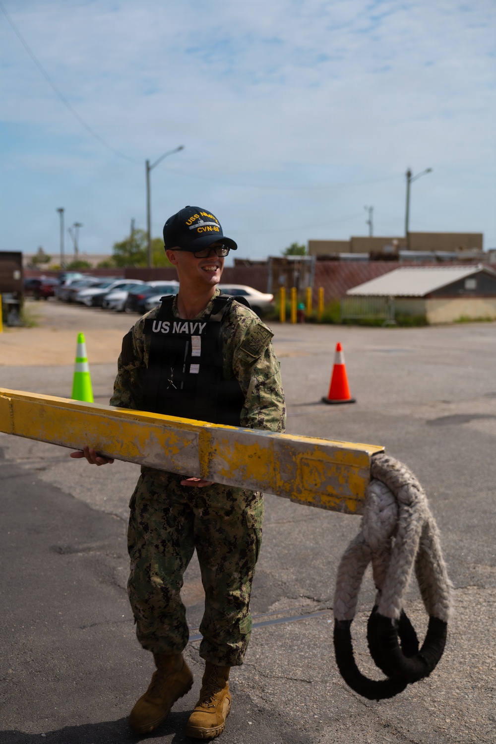 A US Navy sailor in NWU Type III camouflage uniform and tactical vest is holding a yellow painted metal barrier arm at what appears to be a military installation gate or checkpoint. The sailor is wearing a USS Nimitz CVN-68 ball cap and smiling. Traffic cones and a parking area are visible in the background.