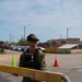 A US Navy sailor in NWU Type III camouflage uniform and tactical vest is holding a yellow painted metal barrier arm at what appears to be a military installation gate or checkpoint. The sailor is wearing a USS Nimitz CVN-68 ball cap and smiling. Traffic cones and a parking area are visible in the background.