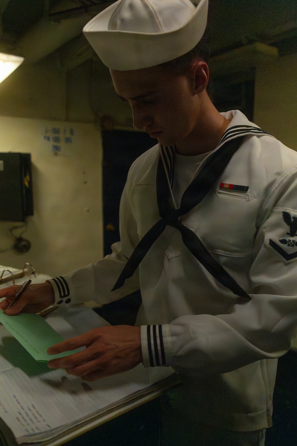 A young U.S. Navy enlisted sailor in dress white uniform is reviewing or signing documents on a clipboard. He is wearing the traditional 'Crackerjack' dress white uniform with a black neckerchief, white Dixie cup hat, and visible rating badge and service ribbon on his chest. The setting appears to be an interior shipboard or shore facility space.