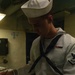 A young U.S. Navy enlisted sailor in dress white uniform is reviewing or signing documents on a clipboard. He is wearing the traditional 'Crackerjack' dress white uniform with a black neckerchief, white Dixie cup hat, and visible rating badge and service ribbon on his chest. The setting appears to be an interior shipboard or shore facility space.