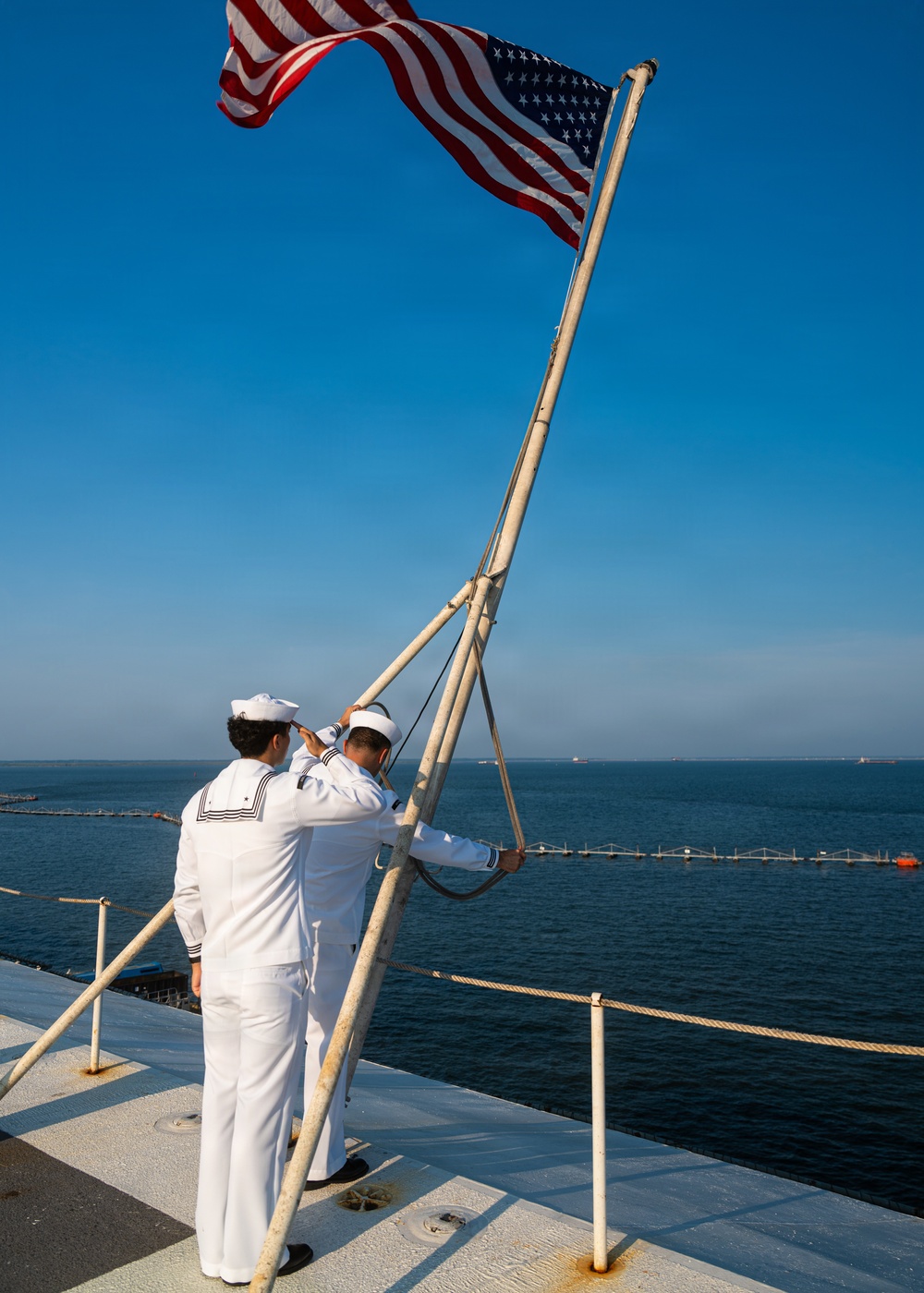 Two U.S. Navy sailors in dress white uniforms are raising or securing the American flag on the stern flagstaff of a naval vessel. The scene is set on the deck of a ship with open ocean and a distant pier or breakwater visible in the background. The clear blue sky and calm sea suggest fair weather conditions.