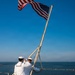 Two U.S. Navy sailors in dress white uniforms are raising or securing the American flag on the stern flagstaff of a naval vessel. The scene is set on the deck of a ship with open ocean and a distant pier or breakwater visible in the background. The clear blue sky and calm sea suggest fair weather conditions.