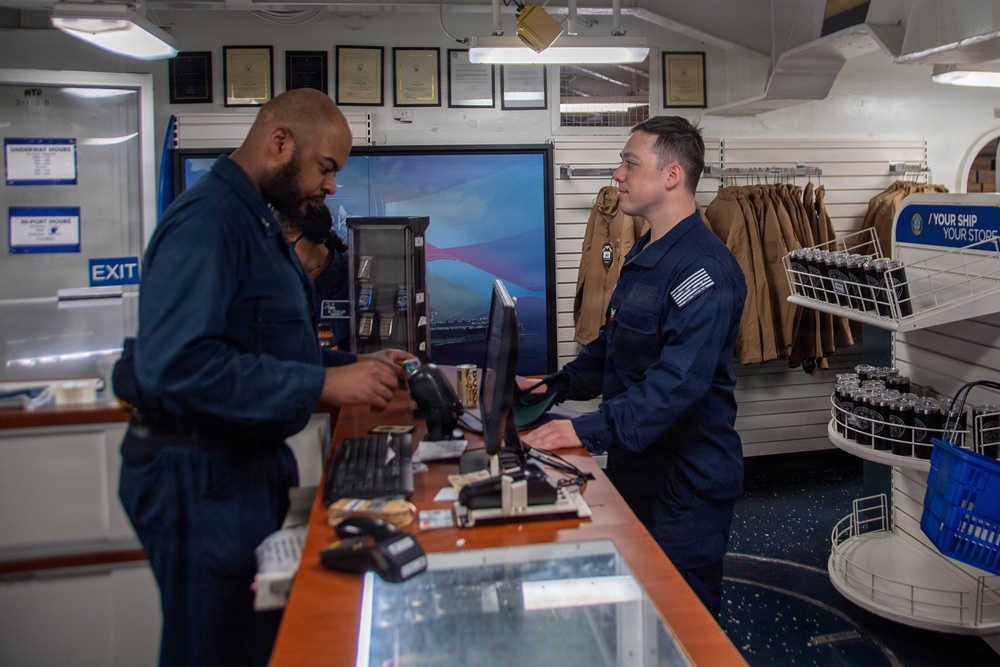 Two personnel in dark blue military uniforms are interacting at a retail counter inside what appears to be a military ship's store. One individual is scanning or handling an item at the point-of-sale terminal while the other stands behind the counter. A sign on the wall reads 'YOUR SHIP YOUR STORE,' confirming this is an onboard military exchange or ship's store.
