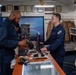 Two personnel in dark blue military uniforms are interacting at a retail counter inside what appears to be a military ship's store. One individual is scanning or handling an item at the point-of-sale terminal while the other stands behind the counter. A sign on the wall reads 'YOUR SHIP YOUR STORE,' confirming this is an onboard military exchange or ship's store.