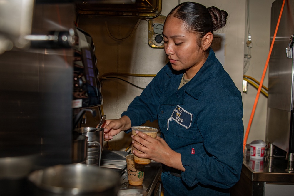 A female sailor in U.S. Navy blue coveralls is preparing a beverage, likely an iced caramel latte, in what appears to be a shipboard galley or coffee station. She is focused on her task, holding a clear plastic cup with a written label visible at the bottom. The setting features industrial piping, cables, and stainless steel equipment consistent with a naval vessel interior.
