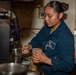 A female sailor in U.S. Navy blue coveralls is preparing a beverage, likely an iced caramel latte, in what appears to be a shipboard galley or coffee station. She is focused on her task, holding a clear plastic cup with a written label visible at the bottom. The setting features industrial piping, cables, and stainless steel equipment consistent with a naval vessel interior.