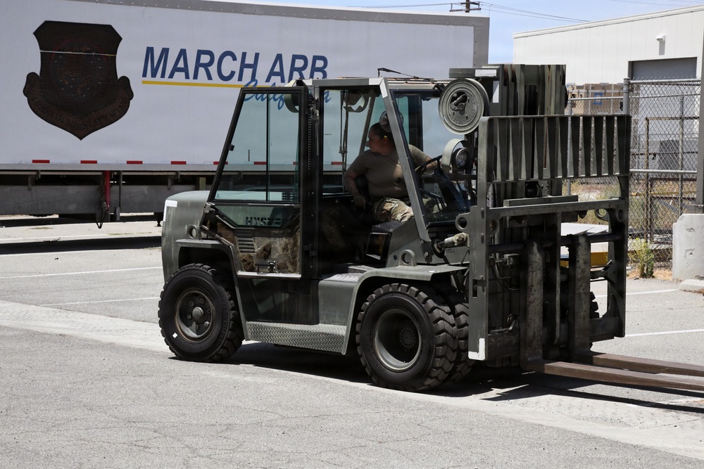 A military service member in camouflage uniform operates an olive-drab/military-green forklift at March Air Reserve Base (March ARB) in California. The forklift's forks are lowered and appear to be carrying or approaching metal rails or beams on the pavement. The setting is an outdoor logistics or cargo handling area on the base.