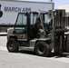 A military service member in camouflage uniform operates an olive-drab/military-green forklift at March Air Reserve Base (March ARB) in California. The forklift's forks are lowered and appear to be carrying or approaching metal rails or beams on the pavement. The setting is an outdoor logistics or cargo handling area on the base.