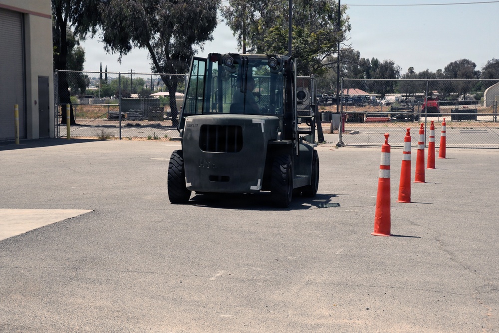 A dark-colored forklift is navigating through a course marked by orange and white traffic cones in an outdoor paved area. The setting appears to be an industrial or military facility, likely used for forklift operator training or certification. The course is set up in a parking lot adjacent to a building with chain-link fencing and trees visible in the background.