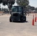 A dark-colored forklift is navigating through a course marked by orange and white traffic cones in an outdoor paved area. The setting appears to be an industrial or military facility, likely used for forklift operator training or certification. The course is set up in a parking lot adjacent to a building with chain-link fencing and trees visible in the background.