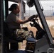 A military service member in camouflage uniform is operating a forklift or heavy equipment vehicle at what appears to be a military installation or base. The individual is seated in the operator's cab, wearing tactical gloves and handling the controls. The background shows chain-link fencing and an open yard area consistent with a military logistics or airfield environment.
