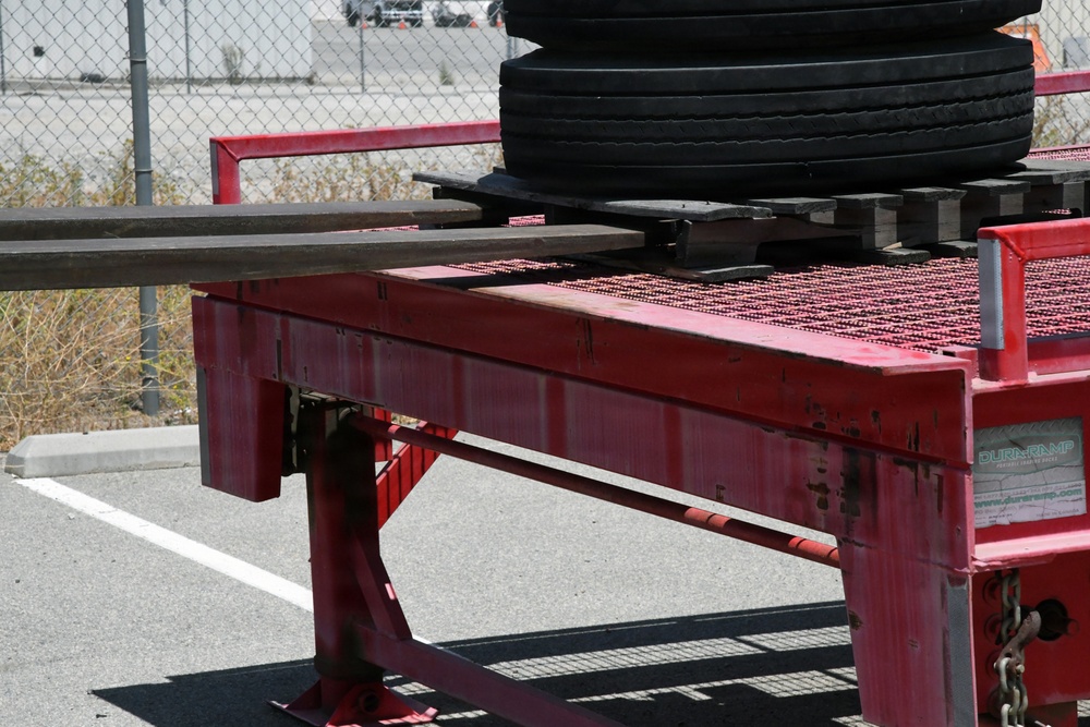 The image shows a red industrial loading ramp or dock leveler, branded 'DuraRamp,' positioned in a parking lot area. Several large truck tires are stacked on a wooden pallet on top of the ramp platform. The equipment appears to be used for loading/unloading heavy cargo, with a chain-link fence and industrial facility visible in the background.