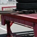 The image shows a red industrial loading ramp or dock leveler, branded 'DuraRamp,' positioned in a parking lot area. Several large truck tires are stacked on a wooden pallet on top of the ramp platform. The equipment appears to be used for loading/unloading heavy cargo, with a chain-link fence and industrial facility visible in the background.