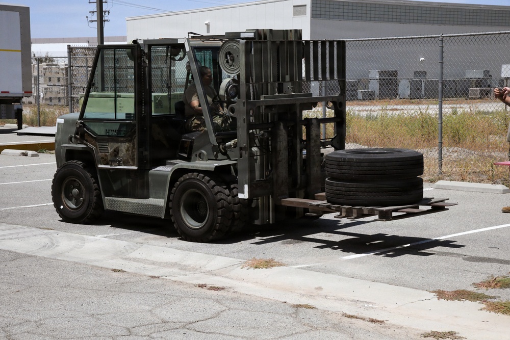 A military-painted Hyster forklift is being operated in a paved parking or staging area, carrying large vehicle tires stacked on a pallet. The operator appears to be wearing military camouflage attire. The setting appears to be a military installation or industrial facility with chain-link fencing and buildings visible in the background.
