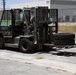 A military-painted Hyster forklift is being operated in a paved parking or staging area, carrying large vehicle tires stacked on a pallet. The operator appears to be wearing military camouflage attire. The setting appears to be a military installation or industrial facility with chain-link fencing and buildings visible in the background.