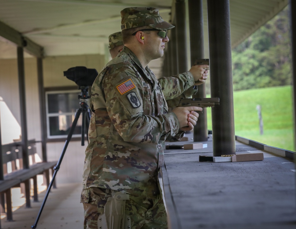 A U.S. Army soldier in MultiCam (OCP) uniform is at an outdoor shooting range, holding a pistol in a firing stance. The soldier is wearing hearing protection (orange earplugs), sunglasses, and a patrol cap, with a U.S. flag patch and unit patch visible on the left sleeve. A second soldier is visible in the background also engaged in pistol shooting.