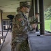 A U.S. Army soldier in MultiCam (OCP) uniform is at an outdoor shooting range, holding a pistol in a firing stance. The soldier is wearing hearing protection (orange earplugs), sunglasses, and a patrol cap, with a U.S. flag patch and unit patch visible on the left sleeve. A second soldier is visible in the background also engaged in pistol shooting.