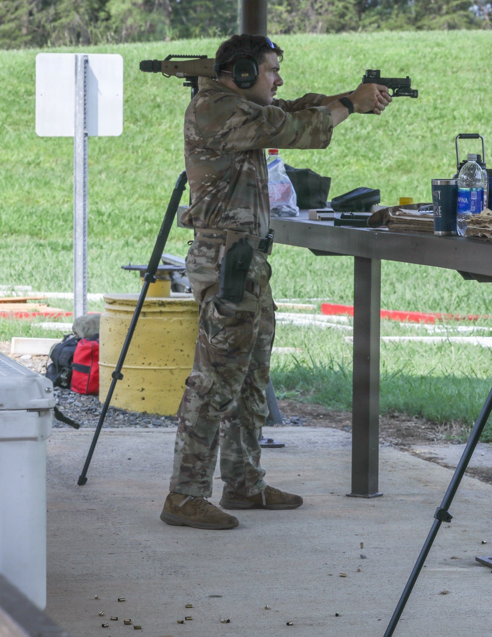 A person wearing multicam/camouflage clothing and hearing protection is actively shooting a pistol at an outdoor shooting range. Spent brass casings are visible on the ground, indicating multiple rounds have been fired. The shooter is in a standing isosceles or modified stance with arms extended, aiming downrange.