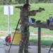 A person wearing multicam/camouflage clothing and hearing protection is actively shooting a pistol at an outdoor shooting range. Spent brass casings are visible on the ground, indicating multiple rounds have been fired. The shooter is in a standing isosceles or modified stance with arms extended, aiming downrange.