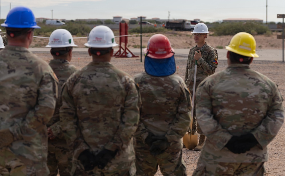 A group of military personnel in camouflage uniforms are gathered outdoors at what appears to be a groundbreaking ceremony on a military installation. One individual facing the group holds a ceremonial shovel and is wearing a white hard hat, while the others wear various colored hard hats including blue, white, red, and yellow. The arid, desert-like terrain and background buildings suggest a southwestern U.S. military base setting.