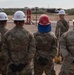 A group of military personnel in camouflage uniforms are gathered outdoors at what appears to be a groundbreaking ceremony on a military installation. One individual facing the group holds a ceremonial shovel and is wearing a white hard hat, while the others wear various colored hard hats including blue, white, red, and yellow. The arid, desert-like terrain and background buildings suggest a southwestern U.S. military base setting.