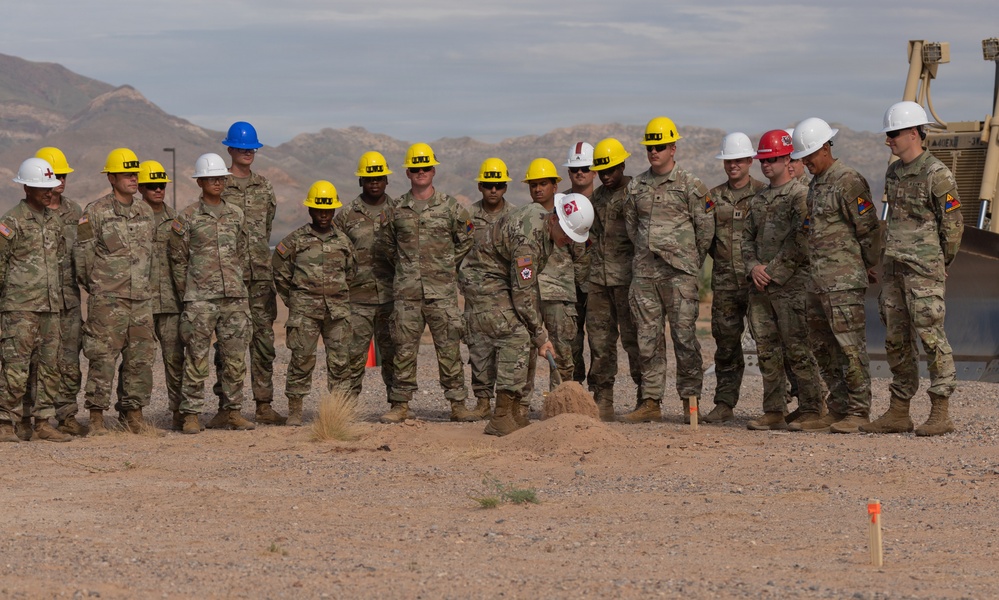 A group of military personnel in camouflage uniforms and hard hats are gathered for what appears to be a groundbreaking ceremony in a desert environment. One individual in the center is using a shovel to break ground while others observe. A construction vehicle is visible in the background, and mountains are visible on the horizon.