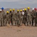 A group of military personnel in camouflage uniforms and hard hats are gathered for what appears to be a groundbreaking ceremony in a desert environment. One individual in the center is using a shovel to break ground while others observe. A construction vehicle is visible in the background, and mountains are visible on the horizon.
