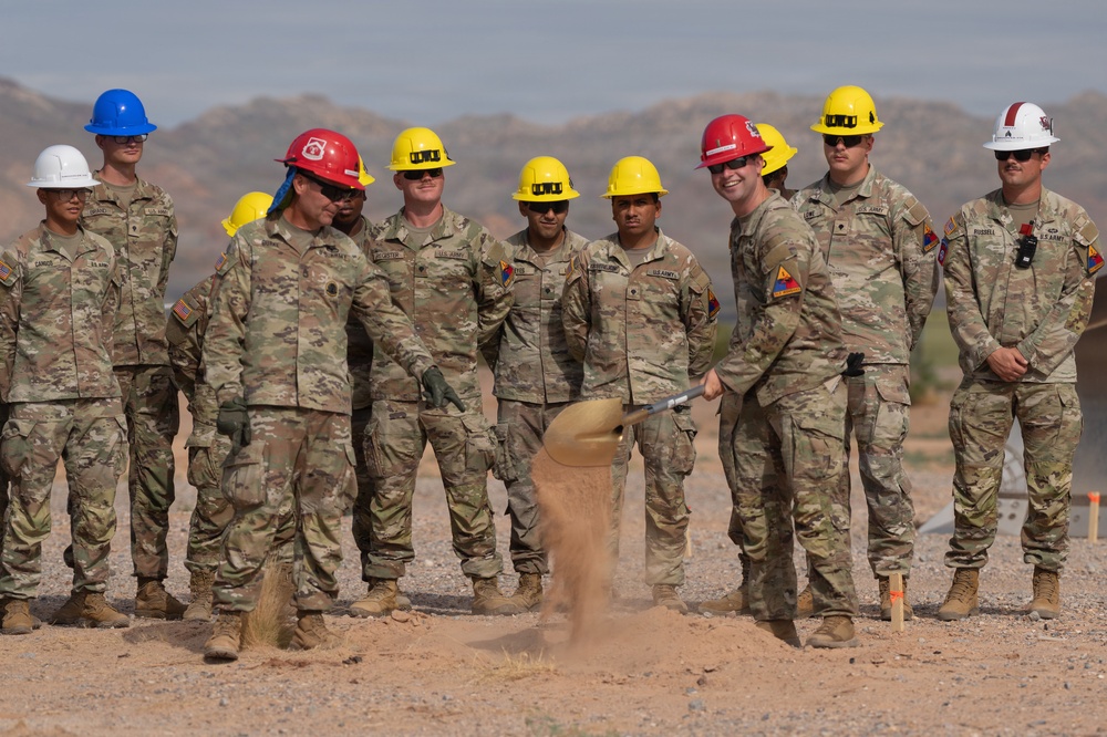 A group of U.S. Army soldiers in camouflage uniforms and colorful hard hats are participating in a groundbreaking ceremony at what appears to be a desert construction site. Two soldiers in the foreground are shoveling dirt, while others stand in a semicircle observing. The arid, mountainous background suggests a southwestern U.S. military installation.