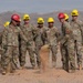 A group of U.S. Army soldiers in camouflage uniforms and colorful hard hats are participating in a groundbreaking ceremony at what appears to be a desert construction site. Two soldiers in the foreground are shoveling dirt, while others stand in a semicircle observing. The arid, mountainous background suggests a southwestern U.S. military installation.