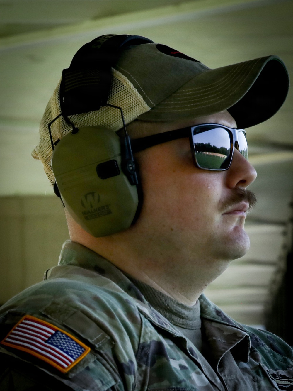 A U.S. Army soldier is photographed in profile view wearing tactical hearing protection and reflective sunglasses. He is dressed in Army Combat Uniform (ACU) with a MultiCam pattern and displays a U.S. flag patch on his left shoulder. The setting appears to be an outdoor shooting range or training facility.