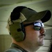 A U.S. Army soldier is photographed in profile view wearing tactical hearing protection and reflective sunglasses. He is dressed in Army Combat Uniform (ACU) with a MultiCam pattern and displays a U.S. flag patch on his left shoulder. The setting appears to be an outdoor shooting range or training facility.