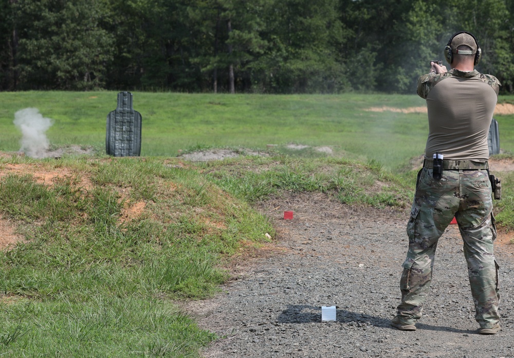 A person in military/tactical camouflage uniform is firing a handgun at a shooting range. Visible gun smoke is present near the target and around the shooter, indicating active firing. The setting is an outdoor range with a grassy field, gravel path, and treeline in the background.
