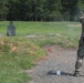 A person in military/tactical camouflage uniform is firing a handgun at a shooting range. Visible gun smoke is present near the target and around the shooter, indicating active firing. The setting is an outdoor range with a grassy field, gravel path, and treeline in the background.