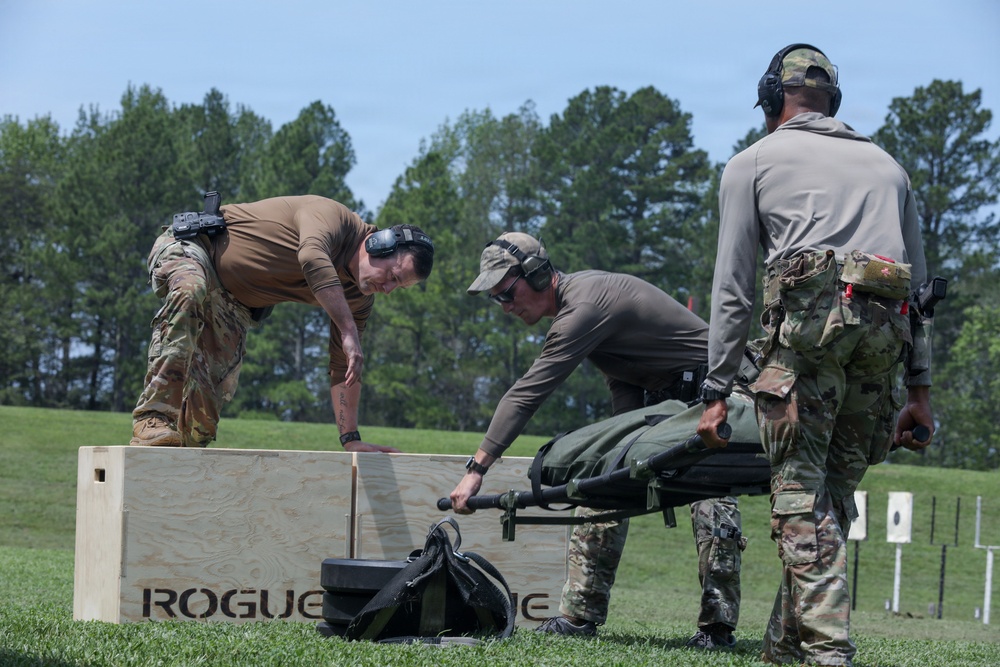 Three military or law enforcement personnel in tactical gear are working together at what appears to be an outdoor shooting range or training facility. They are handling what looks like a litter/stretcher near a large wooden plyo box branded 'Rogue'. The scene suggests a tactical training exercise combining physical fitness elements with operational tasks.