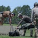 Three military or law enforcement personnel in tactical gear are working together at what appears to be an outdoor shooting range or training facility. They are handling what looks like a litter/stretcher near a large wooden plyo box branded 'Rogue'. The scene suggests a tactical training exercise combining physical fitness elements with operational tasks.