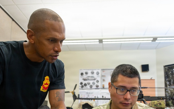Two U.S. Army soldiers are working together at a desk in what appears to be a military training or operations room. One soldier in a black t-shirt with a unit crest patch leans over to review content on a Dell laptop with a seated soldier wearing OCP (Operational Camouflage Pattern) uniform with the name tape 'PABON' and Staff Sergeant rank insignia. A third soldier is visible in the background at another workstation.