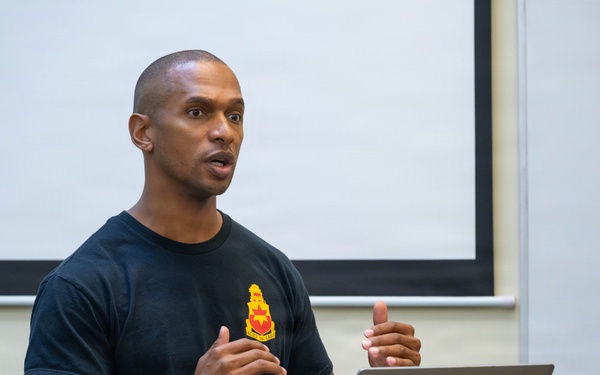 A military service member in a black unit t-shirt and camouflage pants is presenting at a wooden podium with an Apple MacBook laptop open in front of him. He appears to be speaking to an audience in what looks like a classroom or briefing room setting. A projection screen is visible in the background.