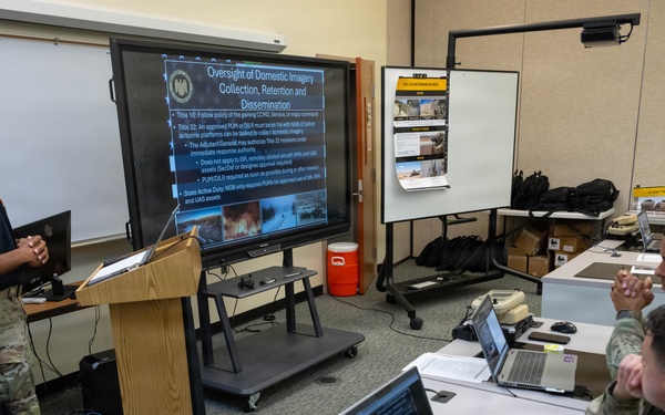 A military briefing or training session is taking place in a classroom setting, with one soldier presenting at a podium beside a large display screen. The presentation slide is titled 'Oversight of Domestic Imagery Collection, Retention and Dissemination.' Three other soldiers are seated at desks with laptops, attentively observing the presentation.
