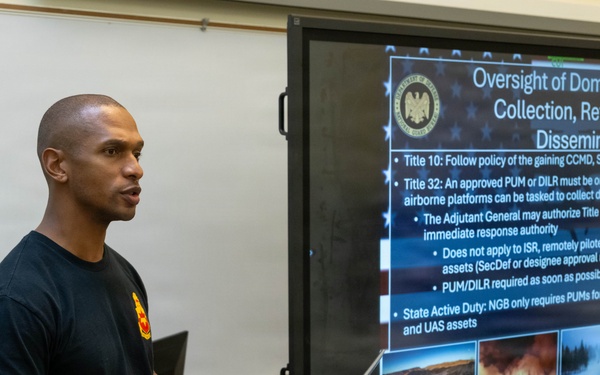 A military service member in uniform is presenting a briefing at a podium in what appears to be a classroom or training facility. The presentation slide displayed on a large screen covers 'Oversight of Domestic Imagery Collection, Retention and Dissemination,' referencing Title 10, Title 32, and State Active Duty regulations. The setting suggests a formal military training or instructional environment.
