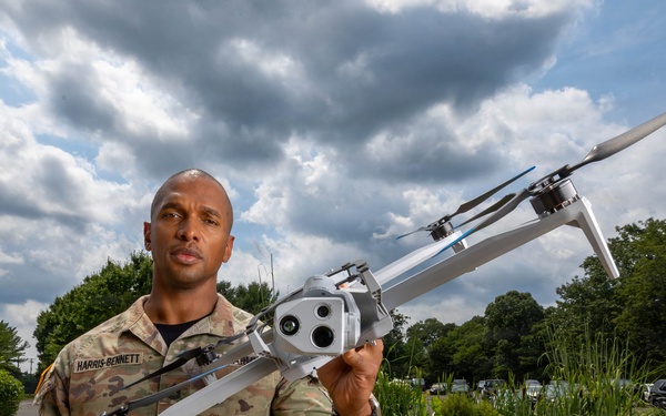 A U.S. Army soldier identified by name tape as 'Harris-Bennett' holds a military unmanned aerial vehicle (UAV/drone) aloft with one hand in an outdoor setting. The drone appears to be a multi-rotor reconnaissance or surveillance UAV equipped with a camera payload. The image appears to be an official or promotional military photograph taken in a landscaped outdoor area.