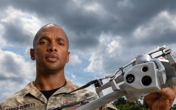A U.S. Army soldier identified by name tape as 'Harris-Bennett' holds a military drone with a multi-lens camera payload at arm's length. The soldier is wearing OCP (Operational Camouflage Pattern) uniform with an 'Instructor' badge visible on the chest. The image appears to be a professional military portrait taken outdoors with a lush green and cloudy sky background.