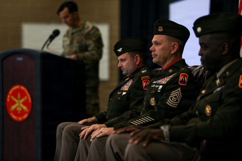 A formal U.S. Army military ceremony is taking place in an indoor venue. Several uniformed soldiers are seated on a stage or dais while one soldier in camouflage uniform speaks at a podium with a microphone. The seated personnel are wearing Army Service Uniforms (green) with various medals, rank insignia, and unit patches visible.