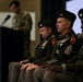 A formal U.S. Army military ceremony is taking place in an indoor venue. Several uniformed soldiers are seated on a stage or dais while one soldier in camouflage uniform speaks at a podium with a microphone. The seated personnel are wearing Army Service Uniforms (green) with various medals, rank insignia, and unit patches visible.